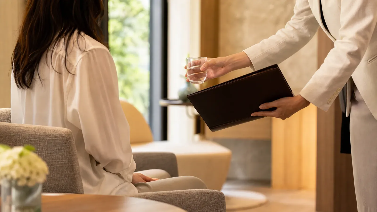 A coordinator offering water and documents to a patient in a quiet private clinic lounge